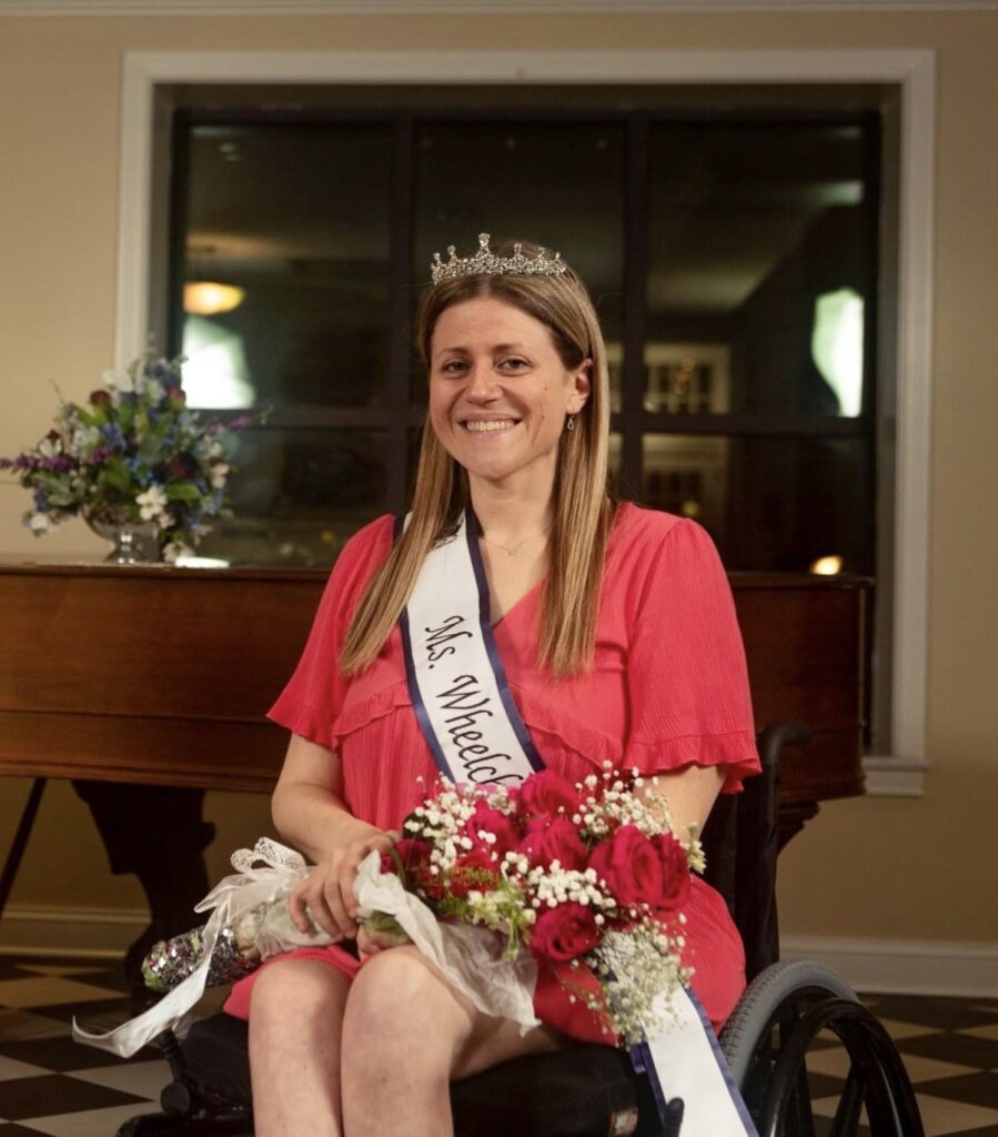Krista is a Caucasian woman with blonde hair, sitting in front of a grand piano at The Mount Vernon Grand Hotel. She is wearing a pink dress, a blue and white Ms. Wheelchair Ohio 2022 sash, and a silver crown atop her head.