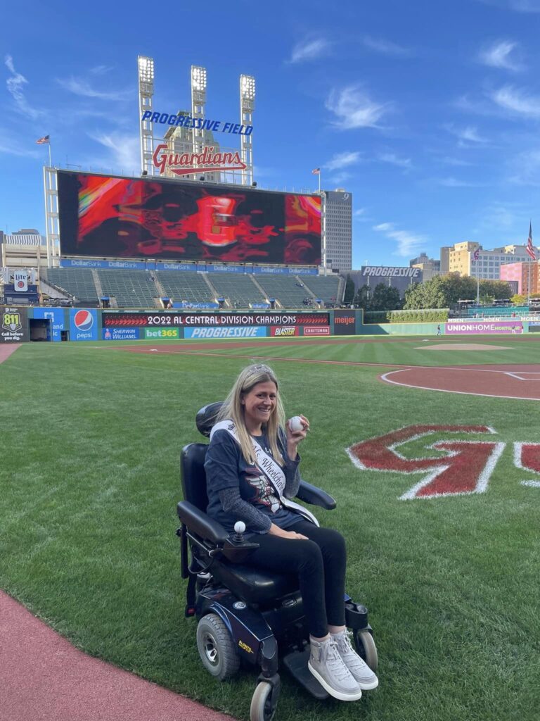 Krista is wearing her crown and sash and holding a baseball. She sits in her powerchair on the Cleveland Guardians' home field in front of a large screen under two signs that read "Progressive Field" and "Guardians."