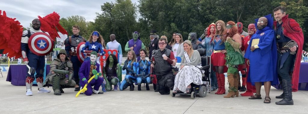 Krista, wearing her sash, crown, and a gray and white snowflake blanket, is photographed in her powerchair surrounded by the group "Superheroes to Kids in Ohio," dressed in hero and villain costumes.