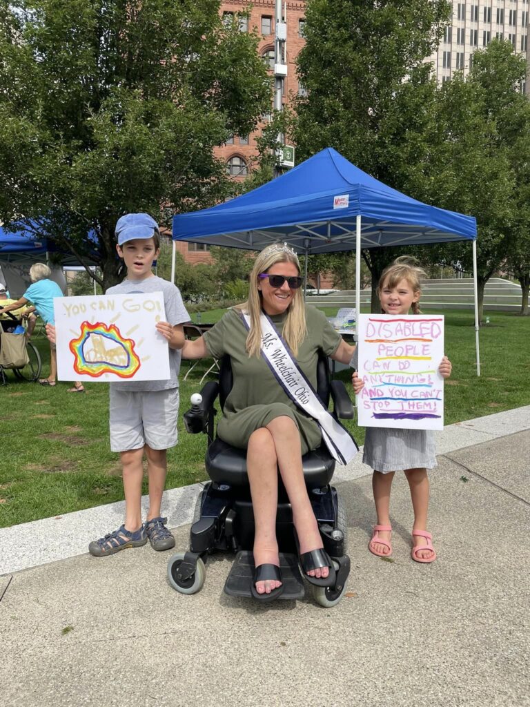 Krista is wearing her sash and crown, sitting outdoors in her powerchair between two children with signs that read, "You can go!," and, "Disabled people can do anything and you can't stop them!"