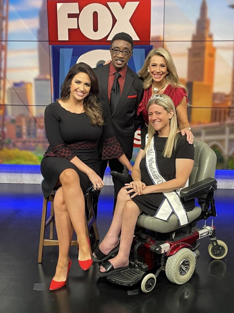 Krista is wearing her sash, crown, and a black dress while sitting in her powerchair surrounded by three Fox 8 News anchors.