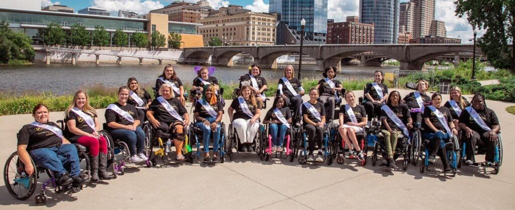 21 women wheelchair users are pictured in front of a grand rapids Michigan waterway below a city skyline. They are all wearing their state sashes, while Ms. Wheelchair America 2021 – Christine Burke wears her Ms. Wheelchair America sash and crown.