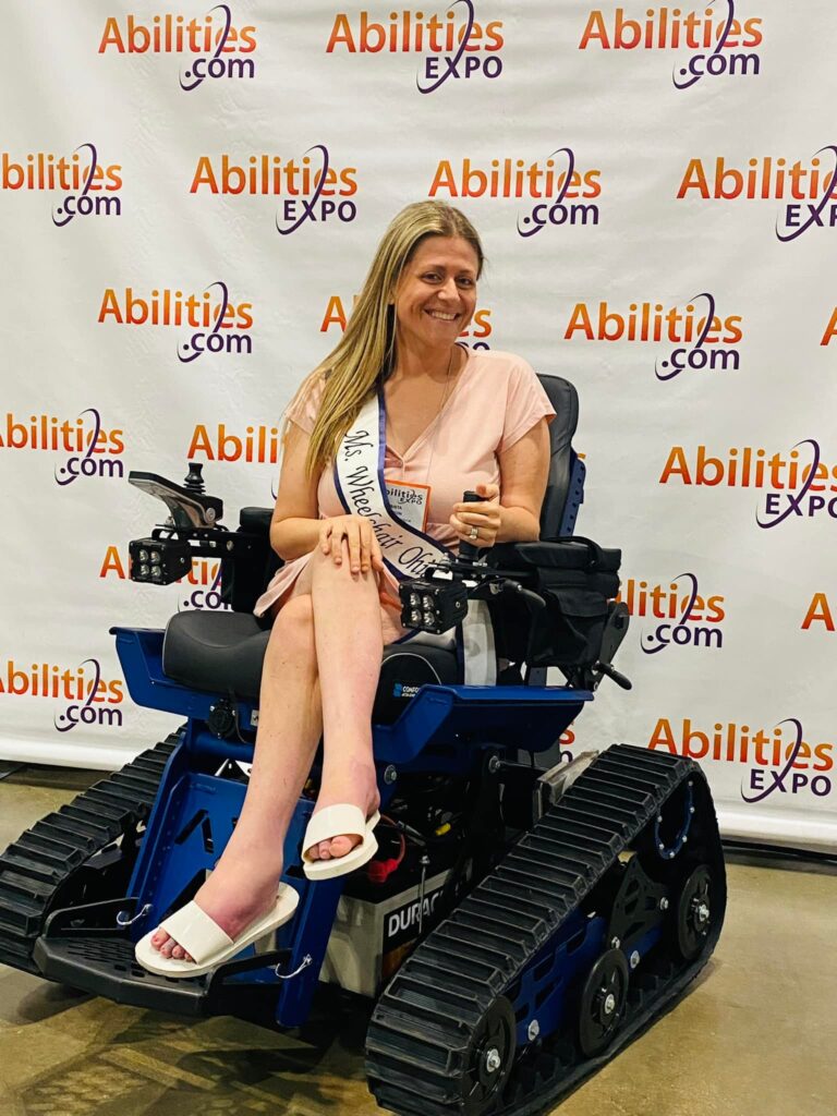 Krista is wearing her sash and sitting in a power chair with utility tracks in front of a backdrop with Abilities Expo's logo and website printed on it.