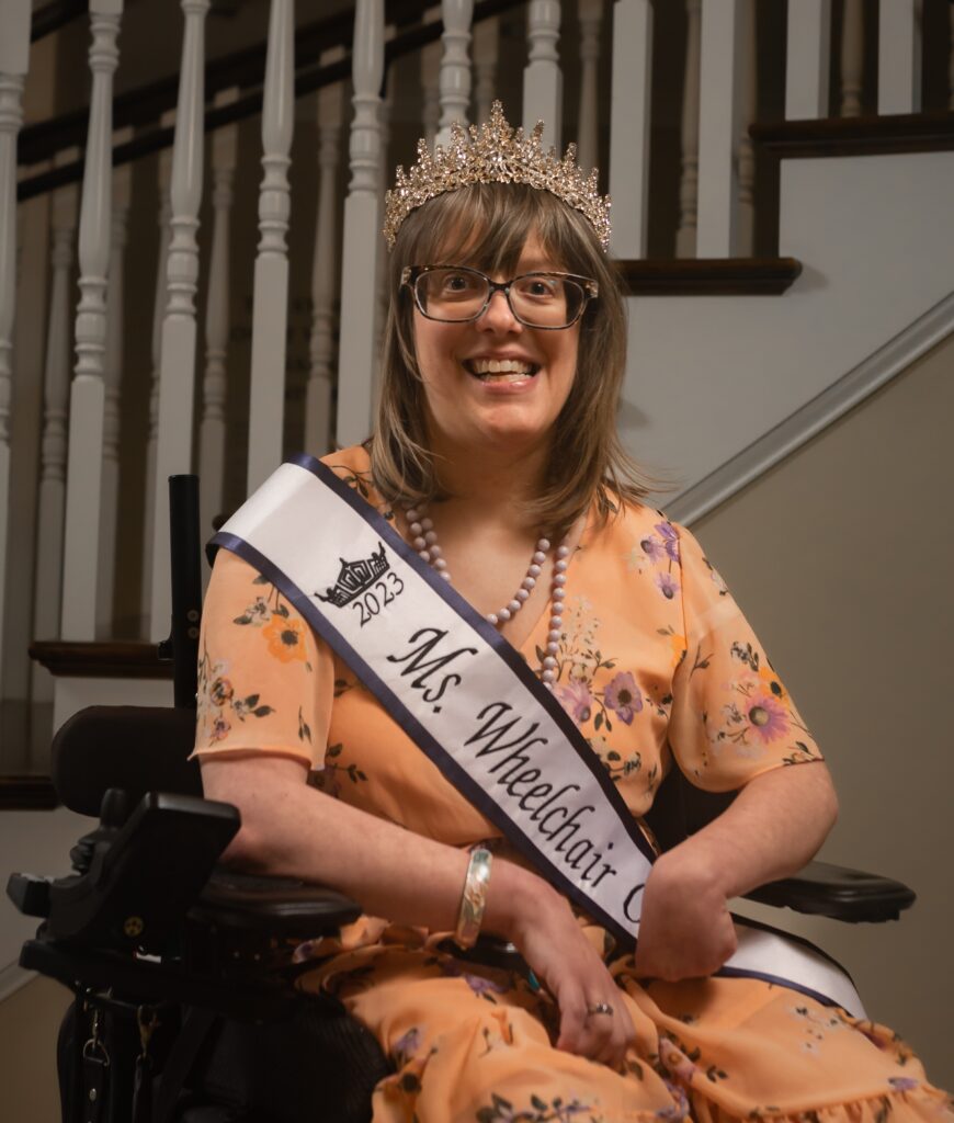 [Image description: Allison is a Caucasian woman with shoulder-length, blonde hair and black glasses. She's wearing an orange floral dress, her Ms. Wheelchair Ohio sash, and a sparkly gold crown as she sits in her powerchair in front of a spiral staircase.]