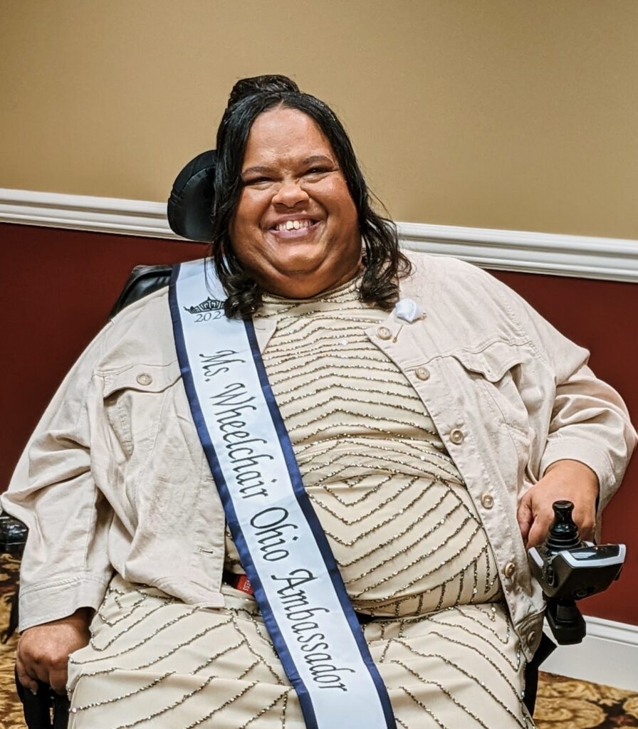 [Image description: Latisha is a Black woman with shoulder-length, black hair. She sits in her powerchair, wearing a tan dress with sparkly, silver accents and her Ms. wheelchair Ohio Ambassador sash.]
