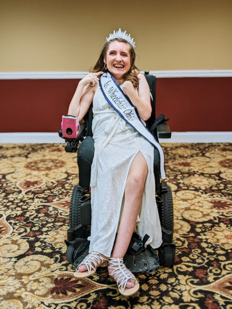[Image description: Gabby is a Caucasian woman with long, brown hair. She sits in her powerchair, wearing a white, sparkly dress, Ms. Wheelchair Ohio sash, and silver, rhinestone crown.]