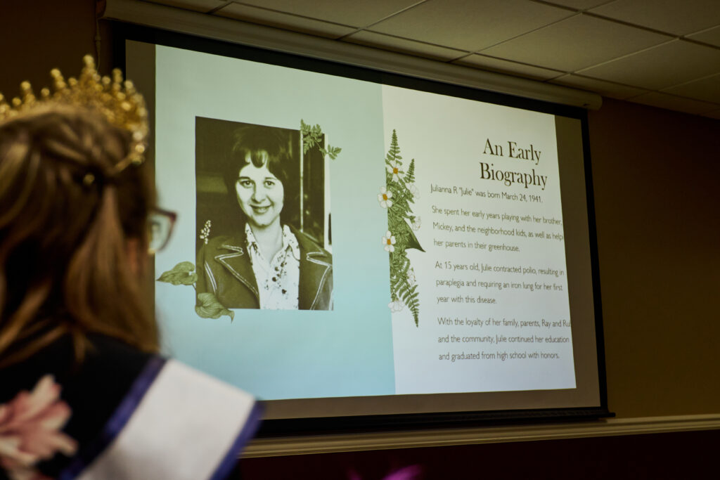 [Image description: A photo of the first Ms. Wheelchair Ohio in 1972, Julie Cochran Rogers, and her early biography are projected on a screen. Out of focus is Allison wearing her crown and sash.]