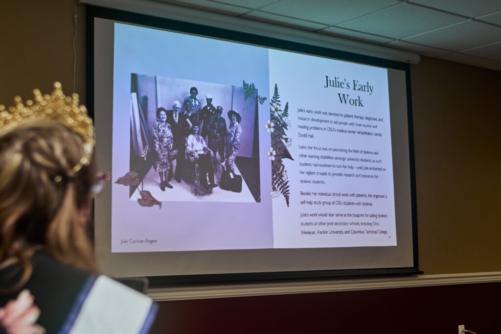 [Image description: A photo of Ms. Wheelchair Ohio 1972, Julie Cochran Rogers surrounded by others and descriptions of her early work are projected on a screen. Out of focus is  Allison wearing her crown and sash.]