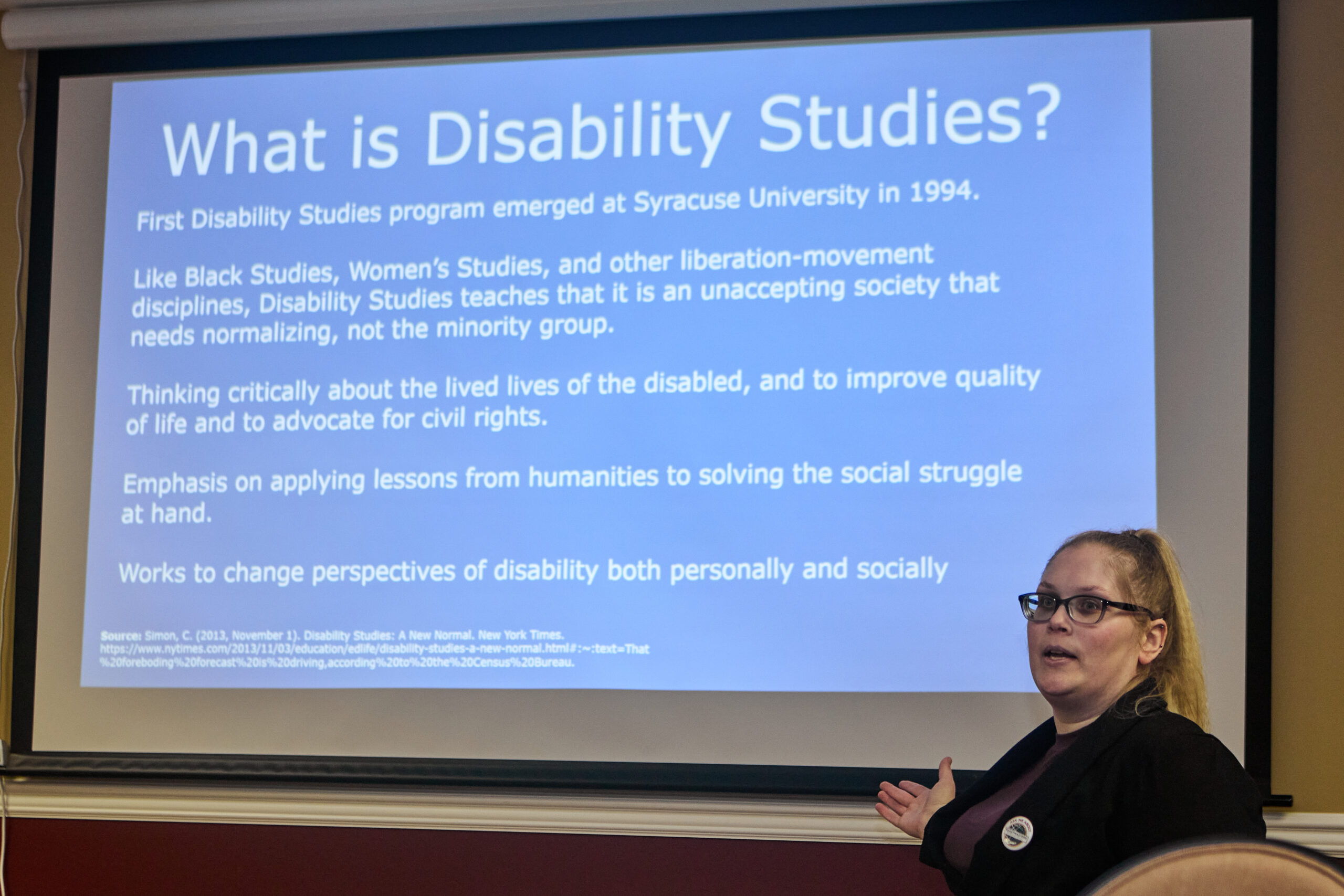 [Image description: Former Ms. Wheelchair Pennsylvania 2018, Barb Zablotney, sitting in front of a screen projecting an explanation of disability studies.]