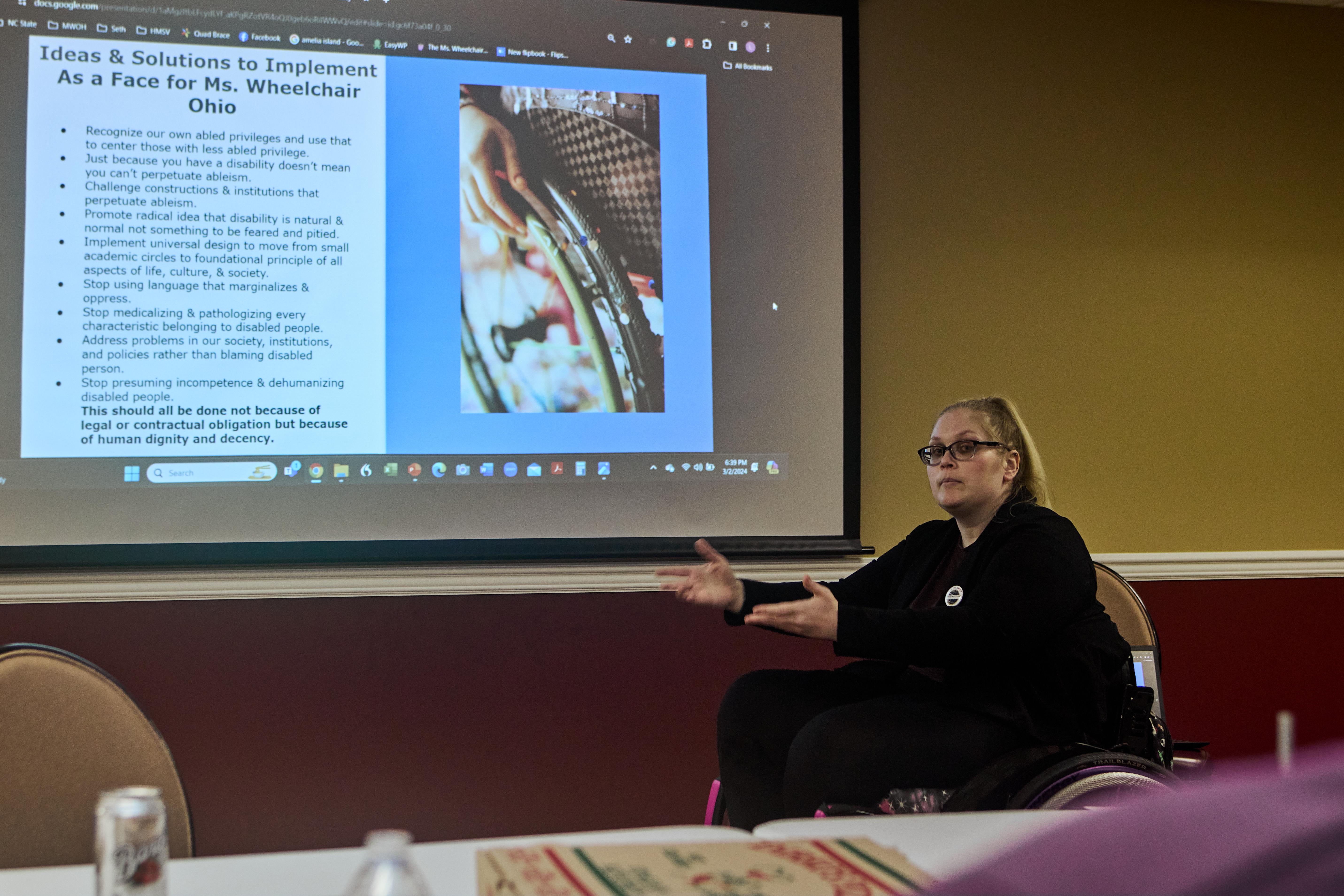 [Image description: Barb sitting in front of a screen projecting ideas to implement disability studies within Ms. Wheelchair Ohio.]