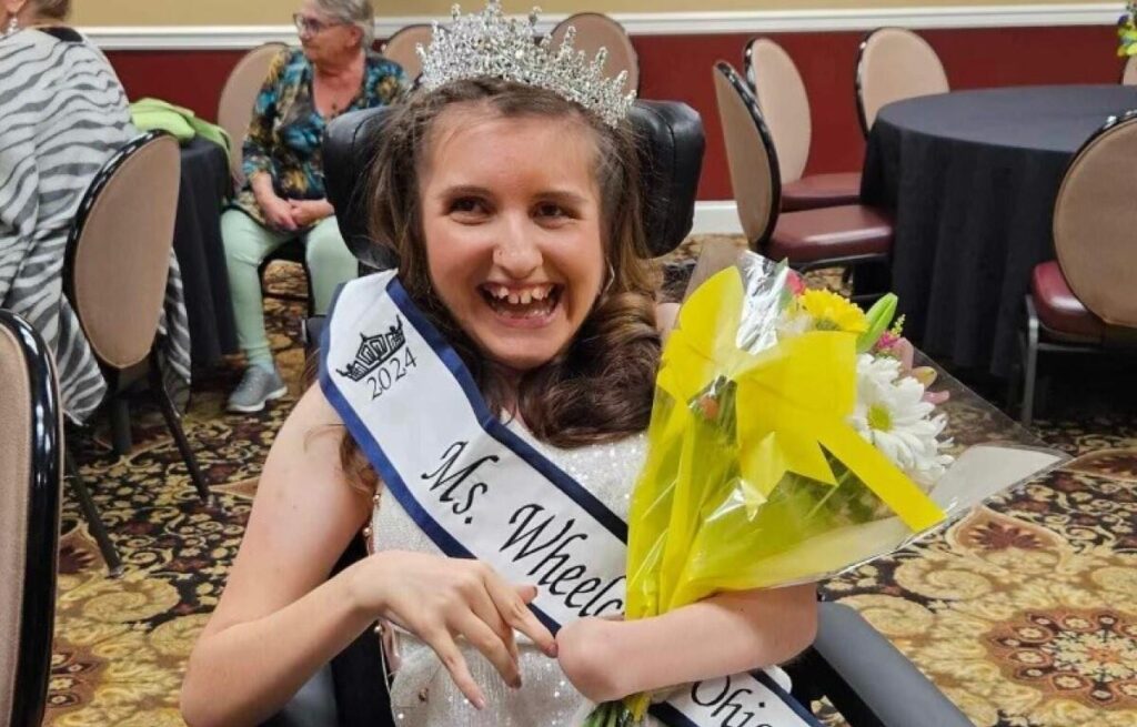 Gabby is smiling big while wearing her crown and Ms. Wheelchair Ohio sash and holding a bouquet of bright colored flowers.