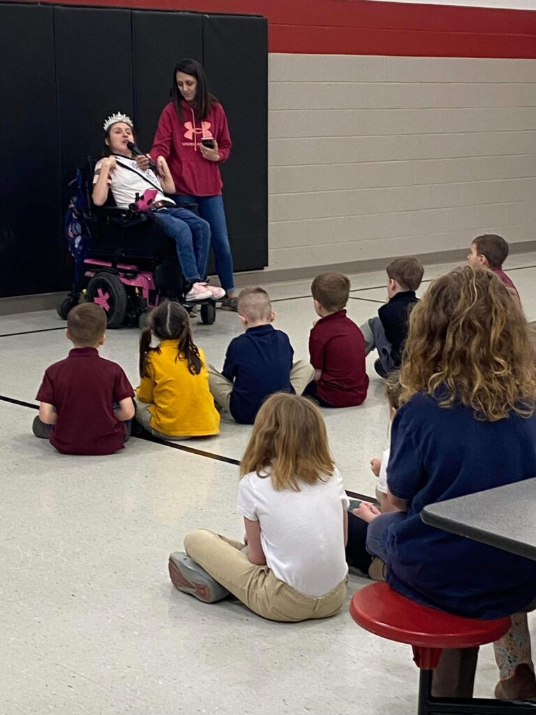 Gabby, wearing her sash and crown, and her mom Kathrine are in front of a group of Sacred Heart students.