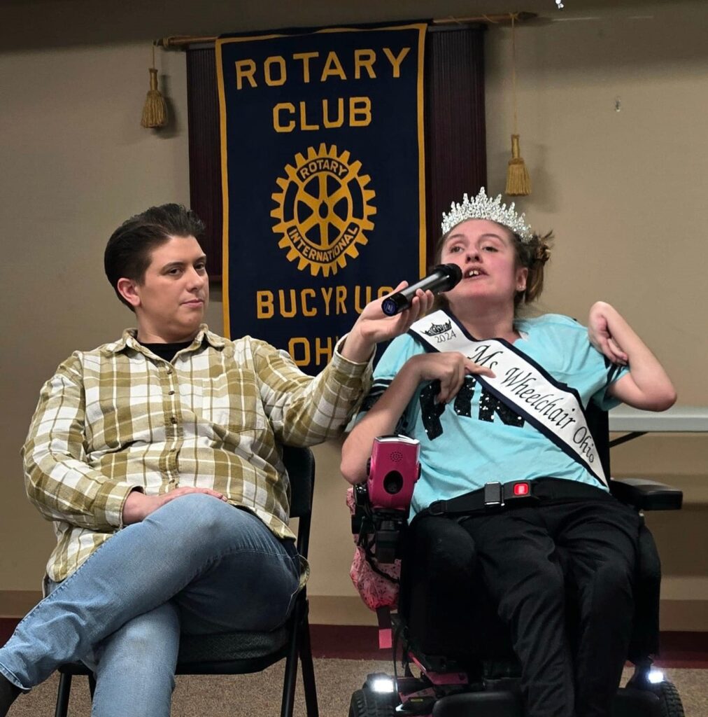 Gabby, wearing her crown and sash, is next to a person holding a microphone. They sit in front of a gold and navy flag that reads, "Rotary Club Bucyrus Ohio."
