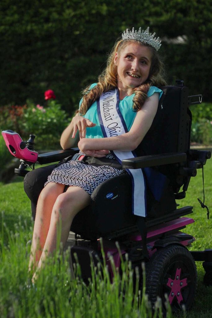 Gabby, wearing her sash and crown, sits in her wheelchair amongst greenery and pink flowers.