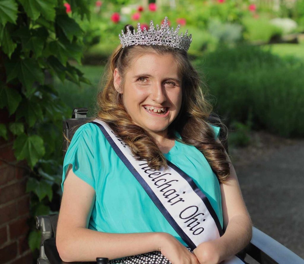 A close-up photo of Gabby shows her wearing her sash and crown in front of greenery and pink flowers.