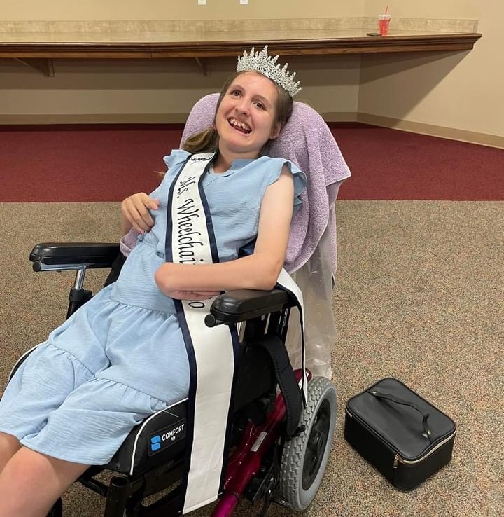 Gabby sits in her wheelchair, wearing her sash, crown, and a big smile.