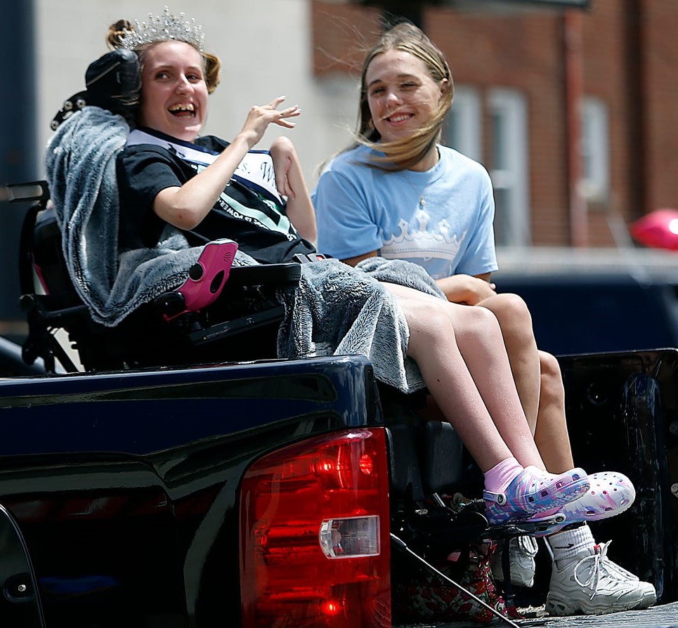 Gabby, wearing her sash and crown, sits in her wheelchair in the back of a pickup truck with her younger sister. Both women are smiling.