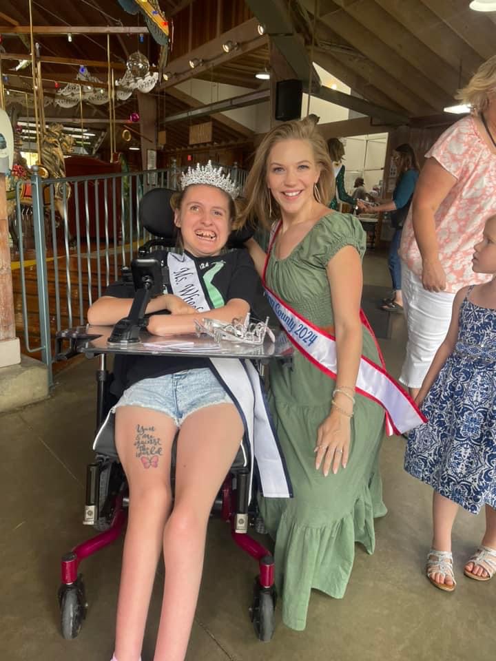 Gabby, wearing her sash and crown, sits in her wheelchair next to another woman wearing a sash. The two women are smiling in front of Mansfield's carousel.