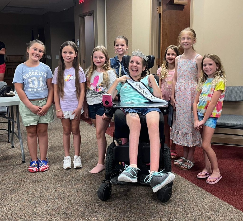 Gabby is wearing her sash and crown and sitting in her powerchair surrounded by a group of young girls. All are wearing big smiles.