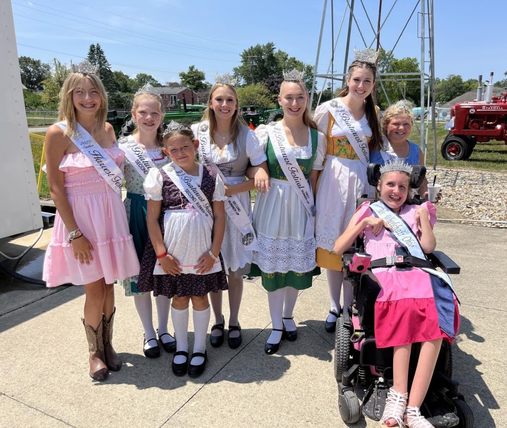 Gabby sits in her powerchair in front of a group of girls wearing dirndls (traditional German dresses). All are wearing sashes, crowns, and big smiles.