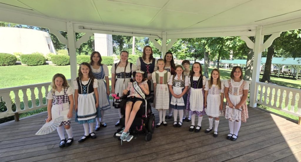 Gabby is wearing her sash and crown and sitting in her powerchair surrounded by girls wearing their dirndls (traditional German dresses).