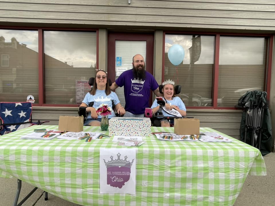 Gabby, wearing her sash and crown, sits next to Seth and Laura behind a table. Atop the table are a green checkered tablecloth, flyers, inclusive coloring pages, and scattered packs of crayons. The Ms. Wheelchair Ohio logo is posted on the front.