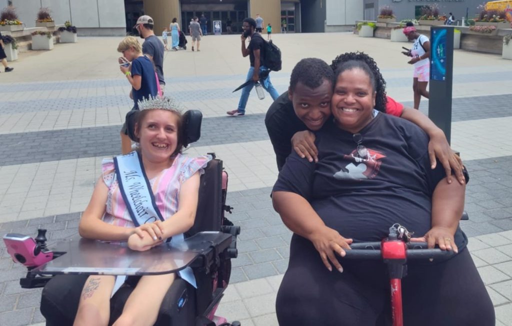 Gabby is wearing her sash and crown and sitting in her powerchair next to a woman on a scooter. A man hugs the scooter-using women from behind.