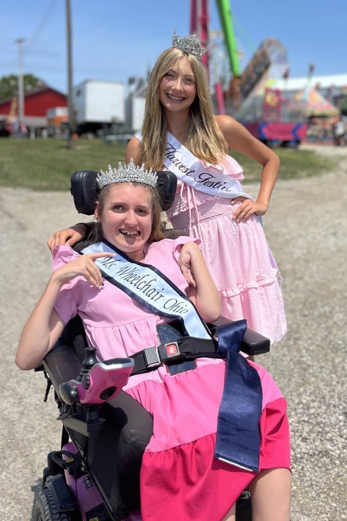 Gabby sits in her power chair next to her younger sister, Eva. Both are wearing their sashes, crowns, and big smiles.