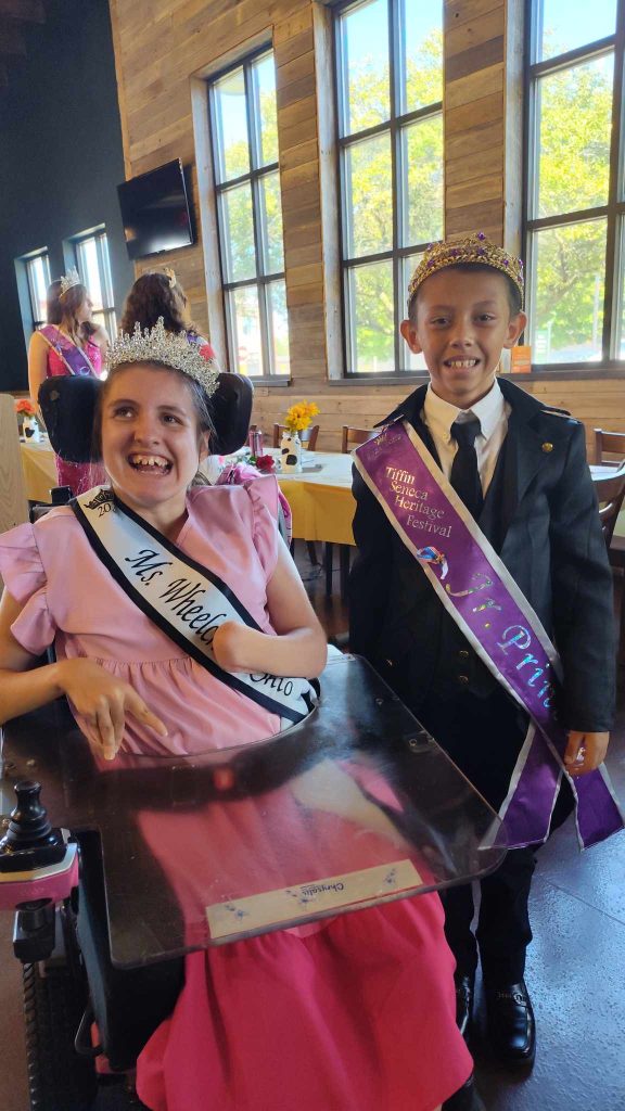 Gabby is wearing her sash and crown sitting in her power chair next to a young boy. The boy is also wearing a sash and crown.