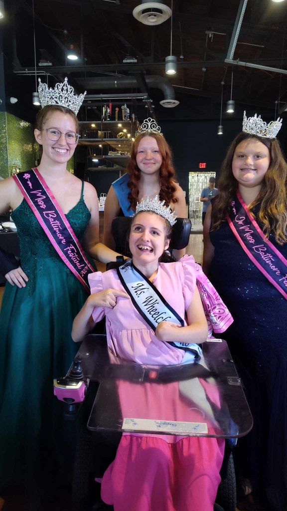 Gabby is wearing her sash and crown sitting in her power chair in front of three girls. The girls are also wearing their crowns and sashes that represent their 2024 titles.