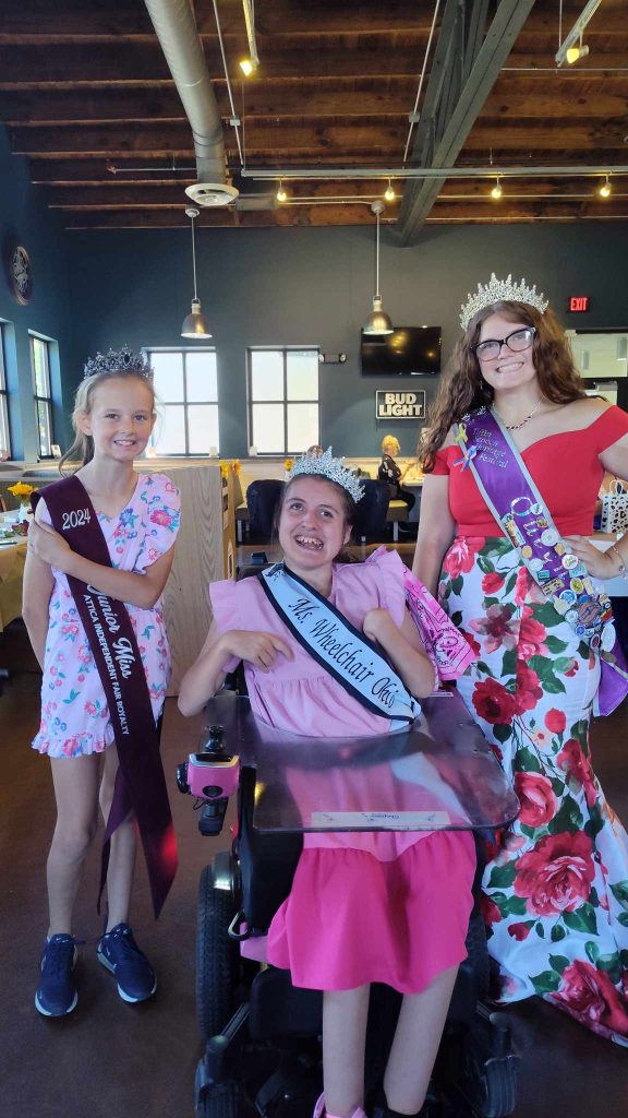 Gabby is wearing her sash and crown sitting in her powerchair between two young girls. The girls are also wearing crowns and sashes that represent their 2024 titles.