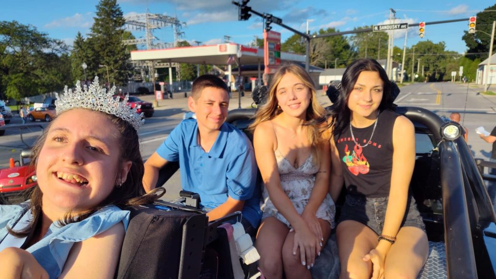 Gabby is wearing her sash and crown, her wheelchair out of frame. Her three siblings sit behind her all in the back of the pickup truck.