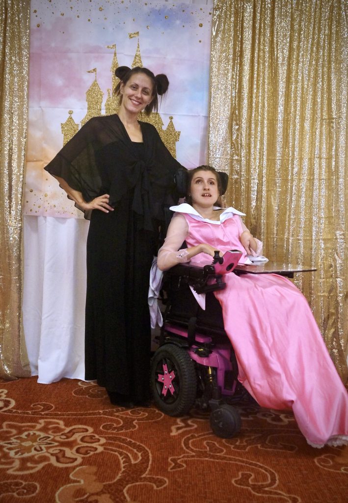 Gabby is wearing a pink gown sitting in her wheelchair, and her mom is wearing a black gown standing behind her. The women are in front of a gold shimmery backed up and the Disney Castle.