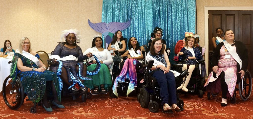 A group of wheelchair-using women in front of a shimmery blue backdrop and colorful mermaid tail. All women are wearing their state sashes.