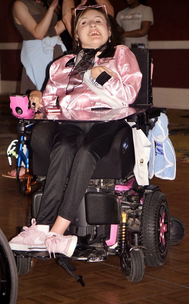 Gabby, sitting in her wheelchair, is wearing a 60s style pink jacket, a black scarf, and pink sunglasses on her head.