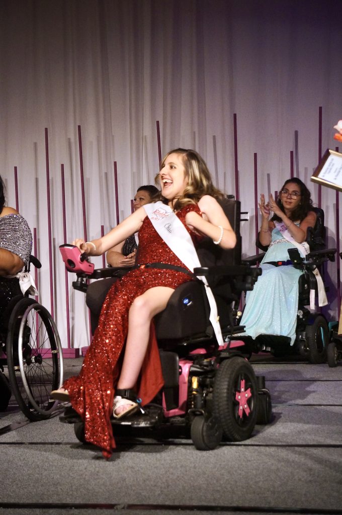 Gabby, sitting in her wheelchair, is wearing a red sequenced dress, her state sash, and a big smile on stage. The women behind her are cheering.