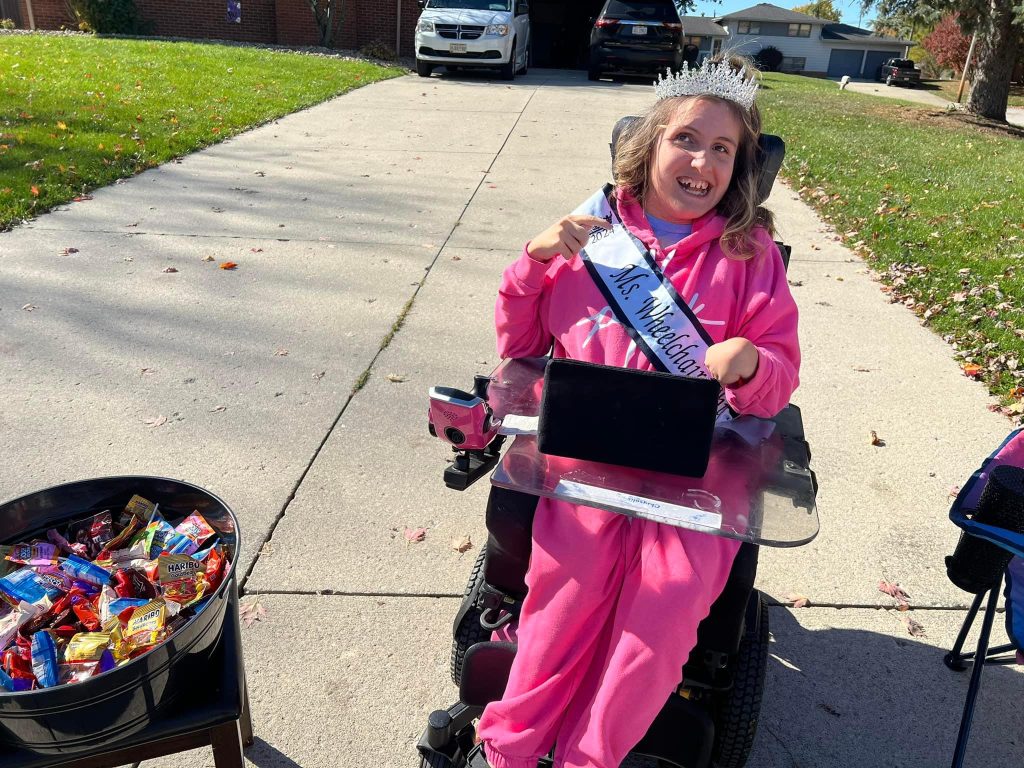 Gabby is wearing a pink bodysuit and her sash and crown. She sits in her powerchair next to a big bowl of candy.