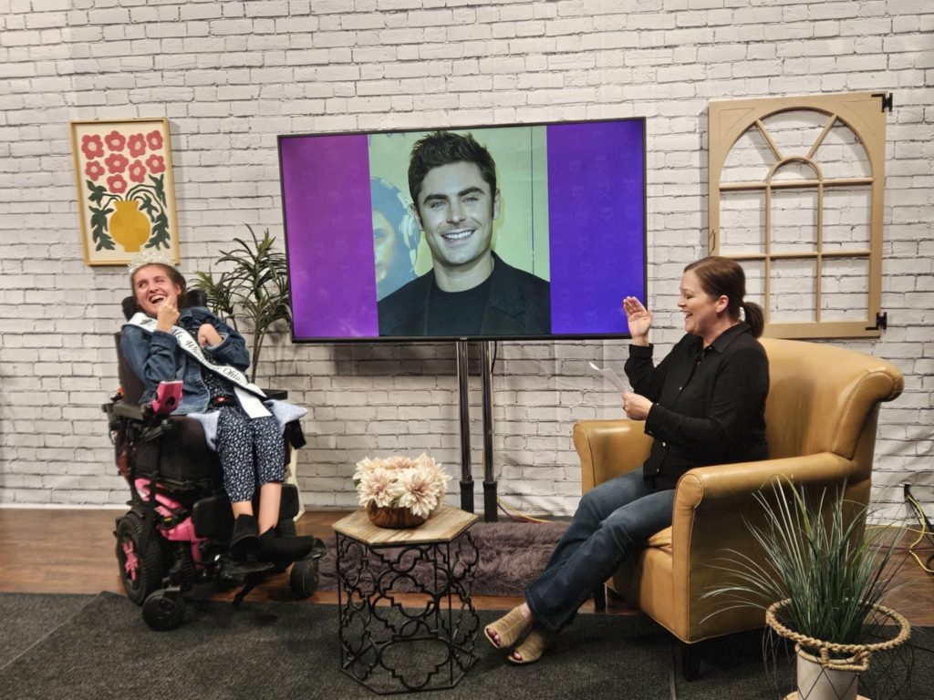 Gabby is wearing her sash and crown and sitting in her powerchair across from Kelby. Both women are laughing in front of a screen projecting a photo of Zac Efron.