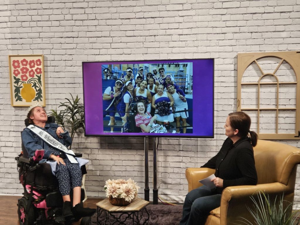 Gabby is wearing her sash and crown and sitting in her powerchair across from Kelby. Both women are looking at a screen projecting a photo of Gabby with Special Olympics cheerleaders.