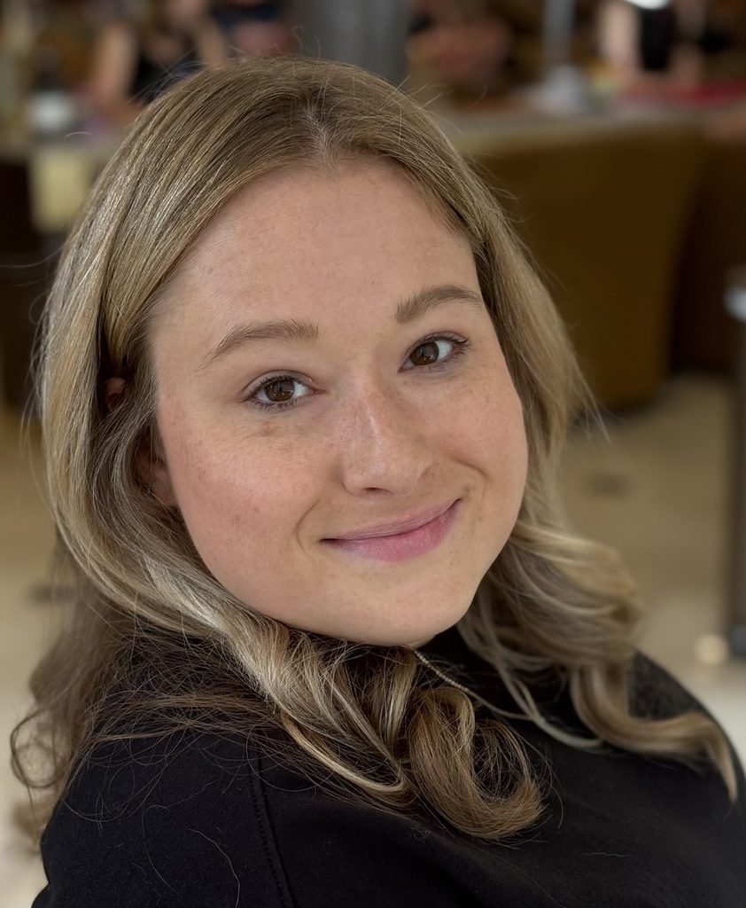 A close-up portrait of a young woman with shoulder-length, light brown hair parted in the middle and softly curled at the ends. She faces the camera with a gentle, closed-mouth smile and a relaxed expression. She is wearing a black top. The photo is taken indoors, with a softly blurred background showing other people and seating, creating a warm, casual setting.
