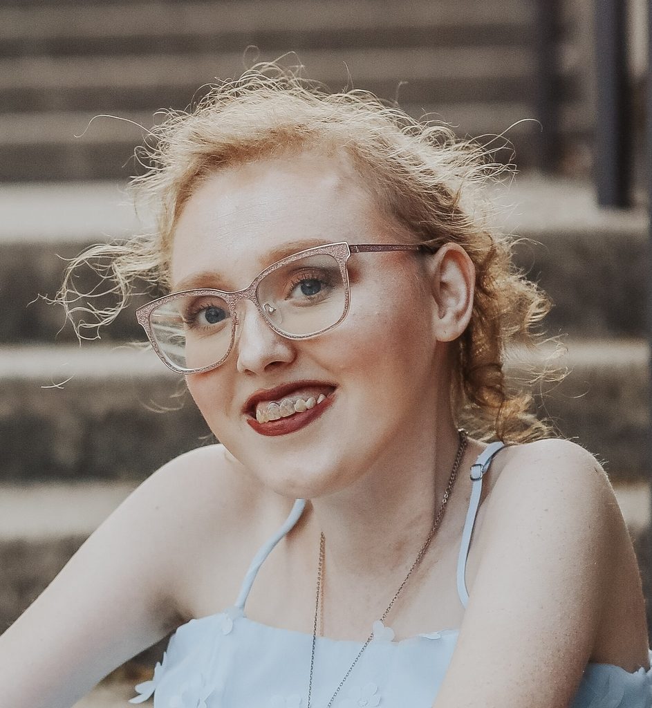 A close-up portrait of a smiling young woman with curly, light-colored hair pulled back loosely. She is wearing sparkling pink-framed glasses and a light blue sleeveless top with small flower details along the neckline. She is also wearing a thin necklace. She faces the camera at an angle, smiling broadly. The background is softly blurred and appears to be an outdoor setting with stone steps or seating behind her.